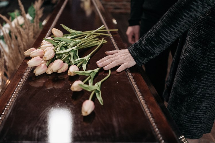 Close-up of a hand resting on a casket with pink tulips placed on top during a funeral service